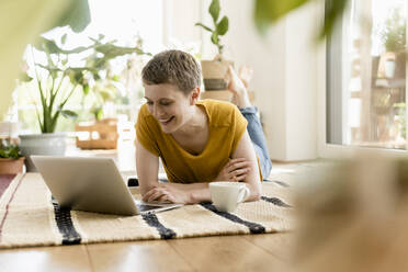 Lächelnde Frau im mittleren Erwachsenenalter, die einen Laptop benutzt, während sie zu Hause auf dem Teppich liegt - UUF21312