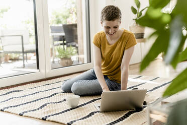 Lächelnde erwachsene Frau mit kurzen Haaren, die einen Laptop benutzt, während sie zu Hause auf einem Teppich sitzt - UUF21314