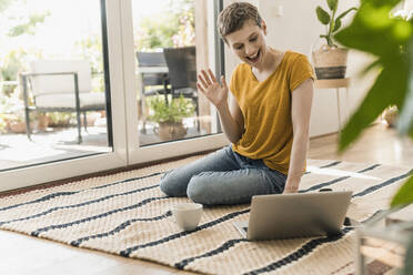 Fröhliche Frau winkt während einer Videokonferenz über einen Laptop auf einem Teppich zu Hause - UUF21315