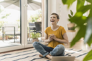 Fröhliche Frau hält lachend ein Getränk in der Hand, während sie mit einem Laptop auf einem Teppich zu Hause sitzt - UUF21316