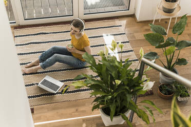 Frau mit Kopfhörern, die ein Getränk in der Hand hält, während sie mit einem Laptop auf einem Teppich zu Hause sitzt - UUF21318