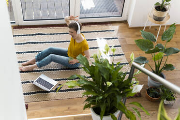 Cheerful woman listening music and dancing while sitting with laptop on carpet at home - UUF21319