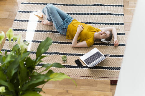 Lächelnde Frau mit geschlossenen Augen hört Musik über Kopfhörer, während sie zu Hause auf dem Teppich liegt - UUF21320