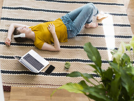 Frau hört Musik über Kopfhörer, während sie mit Laptop und Getränk auf dem Teppich zu Hause liegt - UUF21321