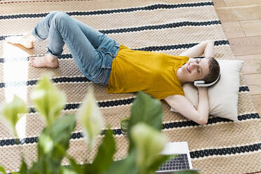 Woman with eyes closed listening music through headphones while lying on carpet at home - UUF21322