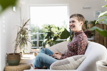 Smiling woman with laptop looking away while sitting on sofa in living room - UUF21323