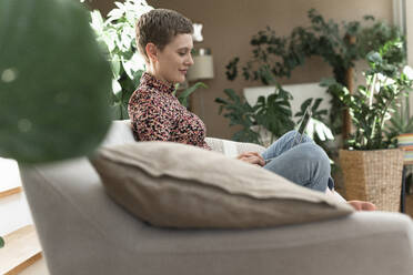 Mittlere erwachsene Frau mit kurzen Haaren, die einen Laptop benutzt, während sie sich auf dem Sofa zu Hause entspannt - UUF21328