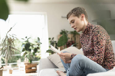 Mid adult woman with short hair reading book while sitting on sofa at home - UUF21330