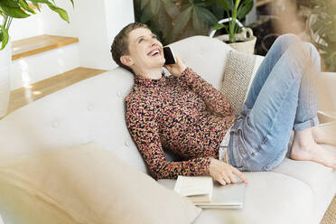 Cheerful mid adult woman talking over smart phone while relaxing on sofa at home - UUF21332
