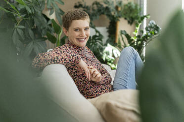 Smiling beautiful woman with short hair sitting on sofa against houseplants in living room - UUF21335