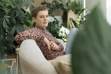 Confident woman with short hair sitting on sofa against houseplants at home - UUF21336