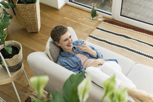 Lächelnde Frau mit kurzen Haaren, die über ein Smartphone spricht, während sie sich auf dem Sofa zu Hause entspannt - UUF21341