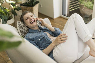 Cheerful woman talking over mobile phone while lying on sofa in living room - UUF21343