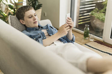 Mid adult woman using mobile phone while resting on sofa in living room - UUF21344