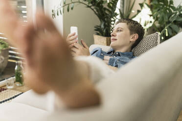Nachdenkliche Frau im mittleren Erwachsenenalter mit Smartphone auf dem Sofa im Wohnzimmer liegend - UUF21345