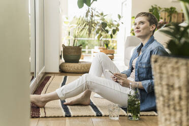 Thoughtful woman with short hair using smart phone while sitting on carpet at home - UUF21347