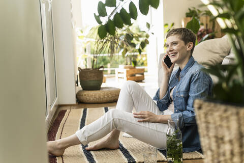 Fröhliche Frau, die über ihr Smartphone spricht, während sie zu Hause auf dem Teppich sitzt, lizenzfreies Stockfoto