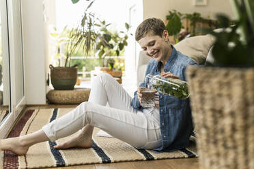 Smiling woman with short hair pouring drink in glass while sitting on carpet at home - UUF21349
