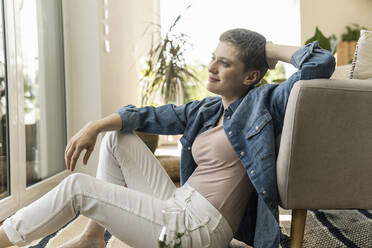 Thoughtful mid adult woman with short hair sitting by armchair at home - UUF21352