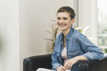 Smiling woman with short hair looking away while sitting on armchair at home - UUF21354