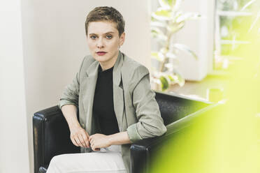 Confident female entrepreneur wearing blazer sitting on armchair at home - UUF21362