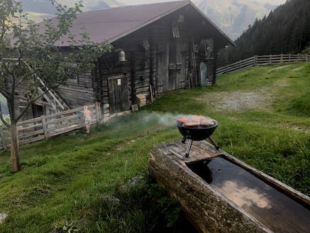 Food cooking on barbecue grill with old cottage in background at Mayrhofen, Tyrol, Austria  - GAF00147