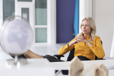 Thoughtful businesswoman with hands clasped sitting on chair in home office - MOEF03192
