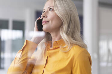 Nahaufnahme einer Geschäftsfrau, die über ein Smartphone im Büro zu Hause spricht, gesehen durch ein Fenster - MOEF03197