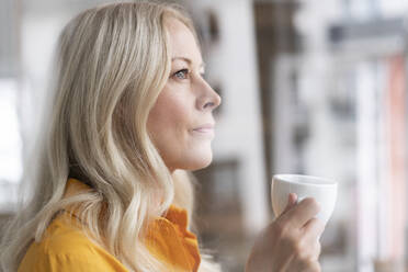 Nahaufnahme einer nachdenklichen Geschäftsfrau, die eine Kaffeetasse im Büro zu Hause hält, gesehen durch ein Fenster - MOEF03200