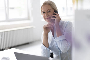 Smiling businesswoman talking over smart phone while sitting at desk in home office - MOEF03212