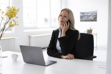 Female entrepreneur with laptop on desk talking over smart phone in home office - MOEF03228