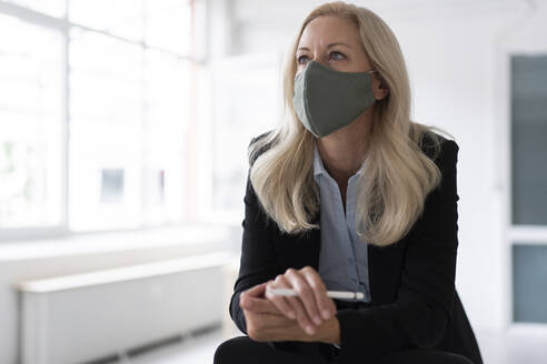 Businesswoman wearing face mask looking away while standing in home office - MOEF03238
