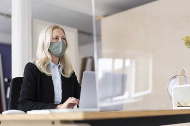 Geschäftsfrau mit Maske, die während der Ausgangssperre einen Laptop auf dem Schreibtisch zu Hause benutzt - MOEF03239