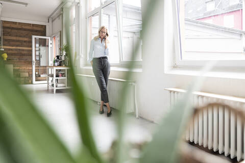Weibliche Fachkraft, die über ein Smartphone spricht, während sie zu Hause am Fenster steht, lizenzfreies Stockfoto