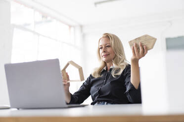 Female entrepreneur holding model home while looking at laptop on desk in home office - MOEF03250