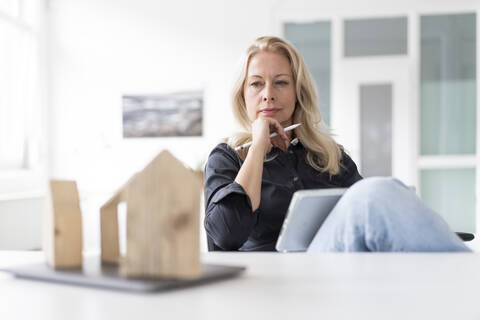 Thoughtful businesswoman with digital tablet looking at model on desk while sitting in home office stock photo