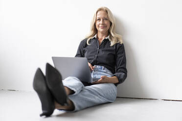 Smiling businesswoman using laptop while sitting on floor against wall in office - MOEF03258