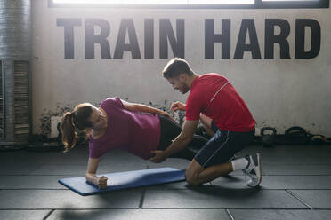 Fitnesslehrer, der einer Sportlerin beim Üben der Plank-Position auf einer Matte im Fitnessstudio hilft - ABZF03289