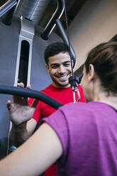 Lächelnder Fitnesstrainer, der einer Frau beim Training mit seitlichen Pull-Down-Gewichten hilft - ABZF03293