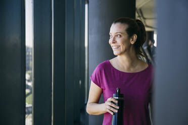 Lächelnde Sportlerin, die eine Wasserflasche hält und durch ein Fenster schaut, während sie im Fitnessstudio steht - ABZF03295