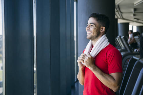 Lächelnder männlicher Sportler, der durch ein Fenster schaut, während er im Fitnessstudio steht - ABZF03300