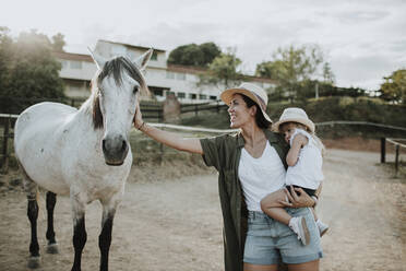 Mother carrying daughter stroking horse while standing on footpath - GMLF00579