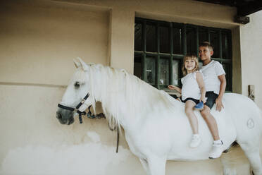 Lächelnde Geschwister sitzen auf einem Pferd an der Wand - GMLF00591