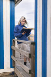 Nachdenkliche junge Frau in warmem Pullover und Rock lehnt an einem Holzzaun mit offenem Buch und liest vor einer weiß und blau gestreiften Wand - ADSF15185
