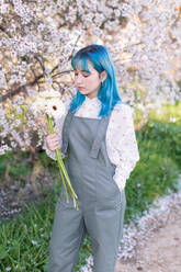 Moderne trendige Frau mit blauem Haar, die einen Strauß frischer Blumen hält und betrachtet, während sie in einem blühenden Frühlingsgarten steht - ADSF15200