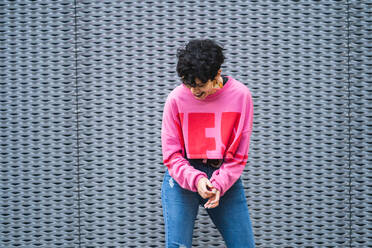 Zuversichtlich fröhlich ethnischen schwarzhaarige Dame in stilvollen rosa Sweatshirt und Jeans stehend mit Hand auf die Taille lächelnd weg vor grauer Wand - ADSF15207