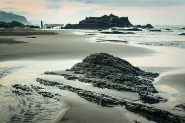 Meereswellen rollen auf nassem Sandstrand mit rauen Felsformationen und entfernte Silhouette von weiblichen Reisenden in Sommerabend in Taiwan - ADSF15211
