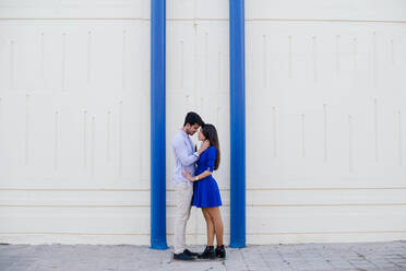 Seitenansicht von liebenden Mann und Frau in eleganter Kleidung küssen auf dem Hintergrund der weißen Wand mit hellen blauen Säulen - ADSF15218