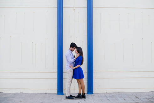 Seitenansicht von liebenden Mann und Frau in eleganter Kleidung küssen auf dem Hintergrund der weißen Wand mit hellen blauen Säulen - ADSF15218