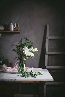 Schöne Glasvase mit Strauß weißer Blumen mit Blättern auf Marmortischplatte neben dunkler Wand - ADSF15220